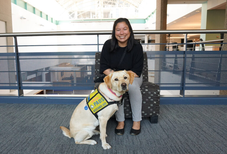 Allison Beardsworth and Wilma who is a yellow Labrador retriever sitting down, posing for the camera. Wilma is wearing a vest saying "PADS assistance dog"
