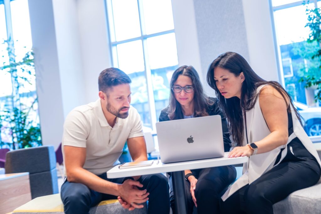 Three individuals sit together in a bright, modern workspace with large windows, collaborating around a silver laptop on a small white table.