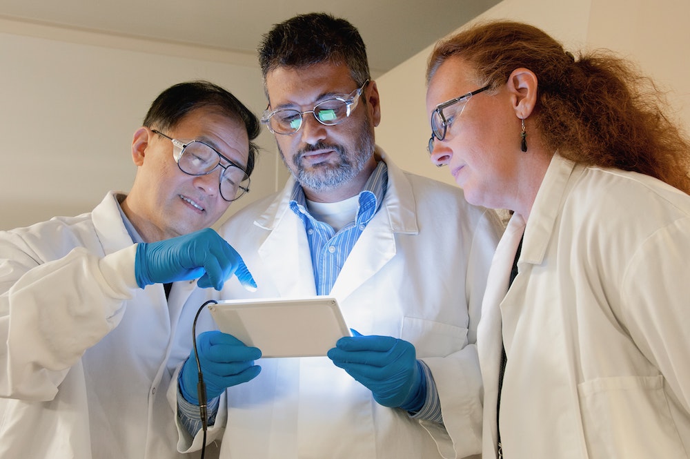 Two male and one female researchers dressed in white lab coats looking at data from a handheld device