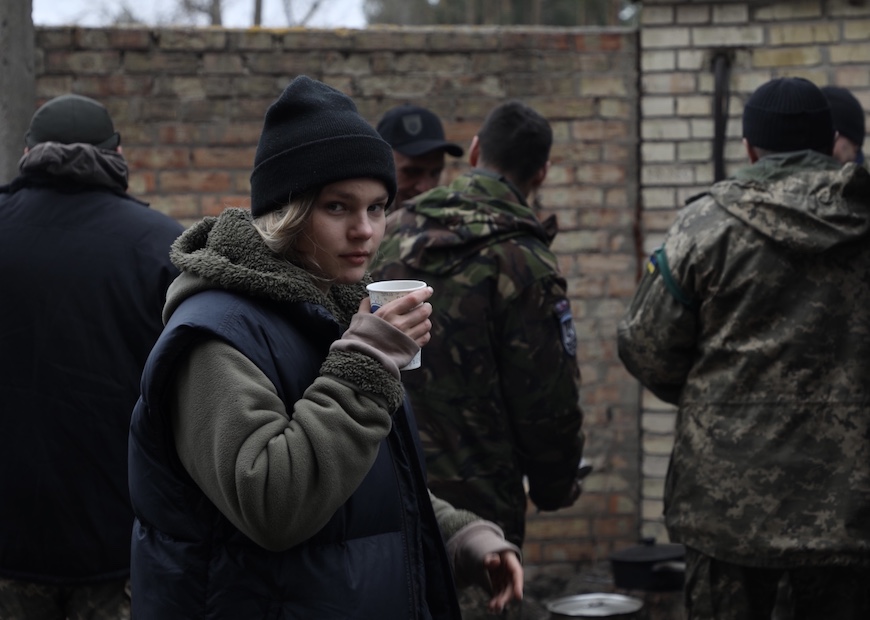 Anastasiia Lapatina looks at camera while drinking coffee with men in camouflage gear in the background