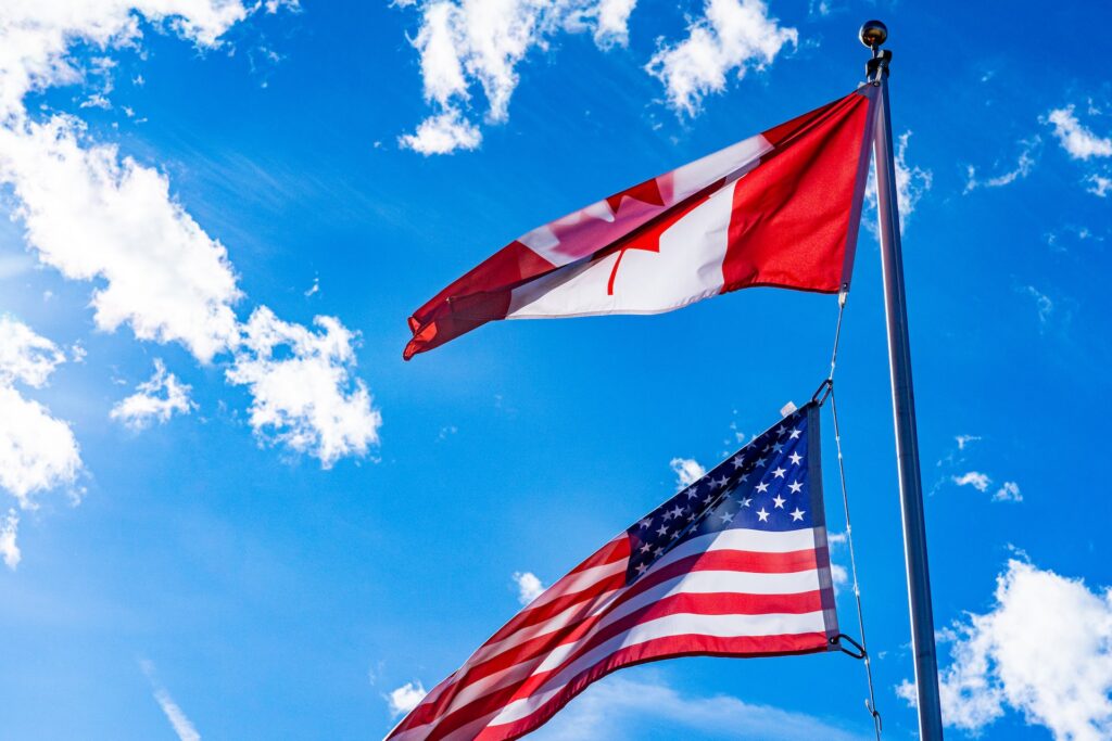 A close-up shot of two flags flying on the same pole. The flag on the bottom is the American flag, recognizable by its red and white stripes and blue field with white stars. The flag on top is the Canadian flag, featuring a red field with a white square in the center bearing a stylized red maple leaf. The clear blue sky in the background provides a beautiful contrast to the colorful flags fluttering in the breeze.