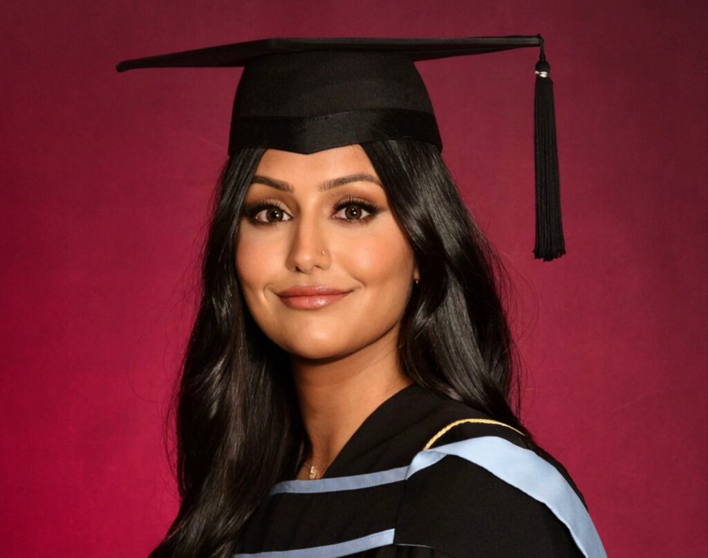 Headshot of Rabiah Dhaliwal, a south Asian young woman, infront of a red background. She's wearing a mortar board and a graduation robe that has a light blue hood on her shoulders.
