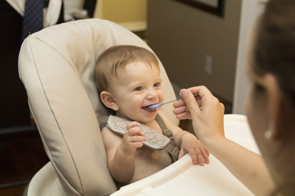 A young infant is sitting in a baby chair with their mouth open as they are being fed food from a small spoon.