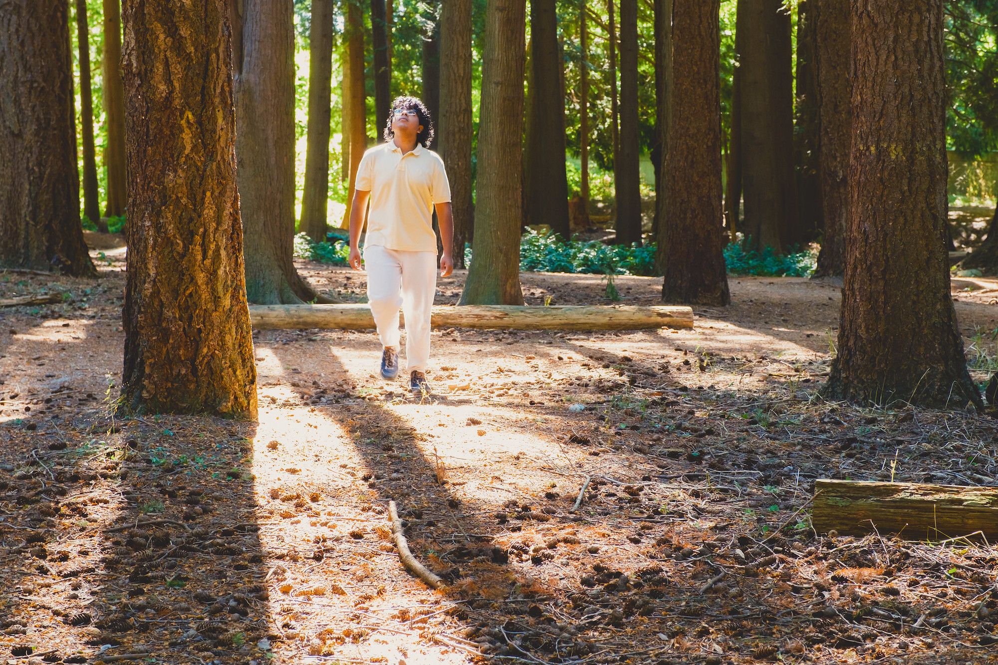A South Asian student dressed in all white walking amongst pine trees on campus.