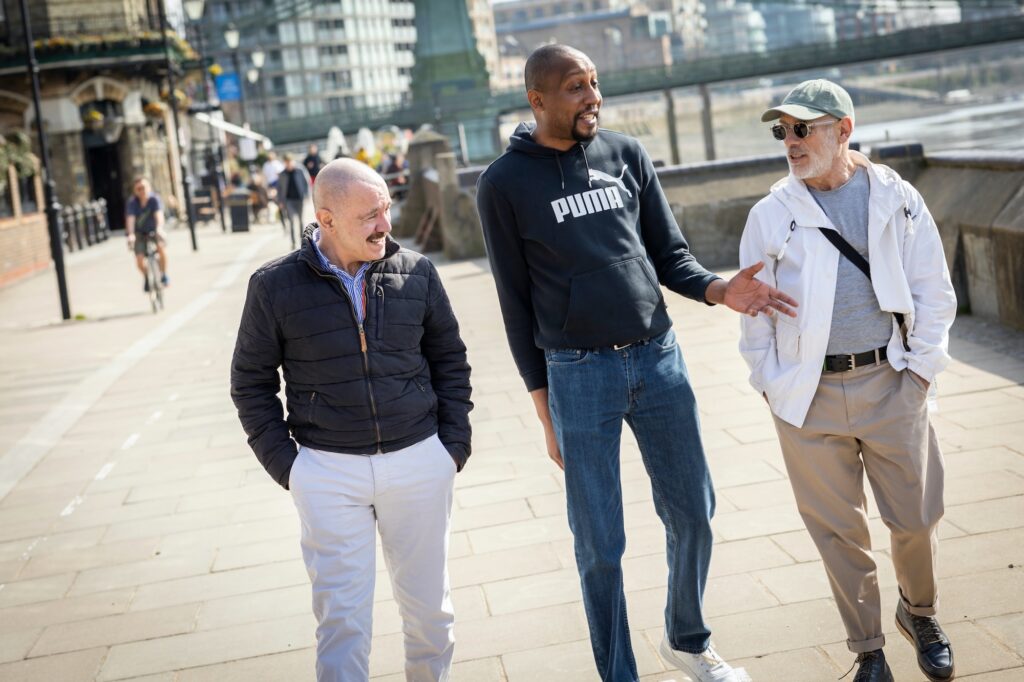 A group of three middle-aged men - two caucasian and one Black - walking together on a boardwalk chatting with each other.