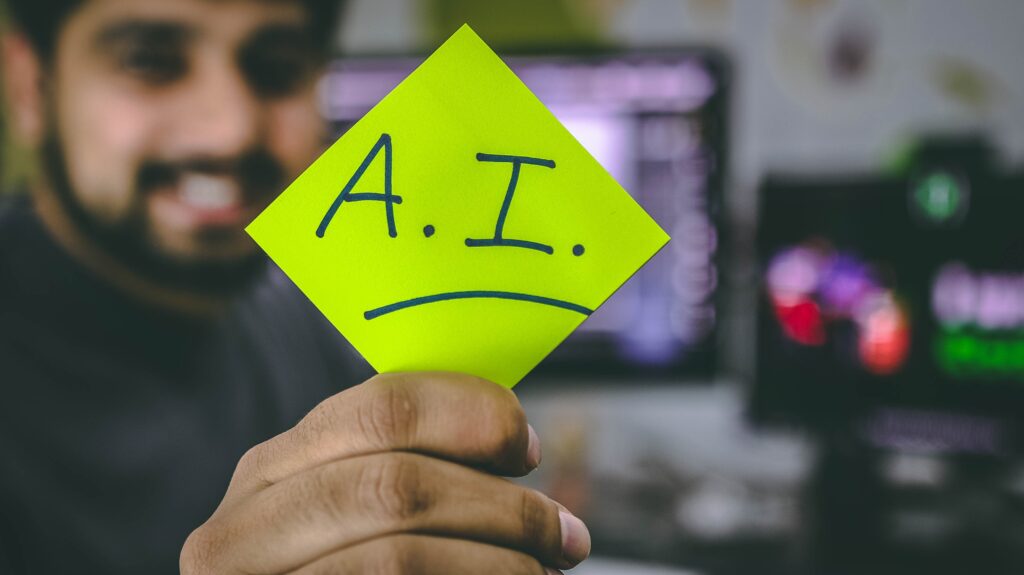 In the foreground, a South Asian man is slightly out of focus, extending his hand forward. In his hand pulled out to the front and in focus, a bright neon yellow post-it note is prominently displayed, bearing the written letters "A.I."