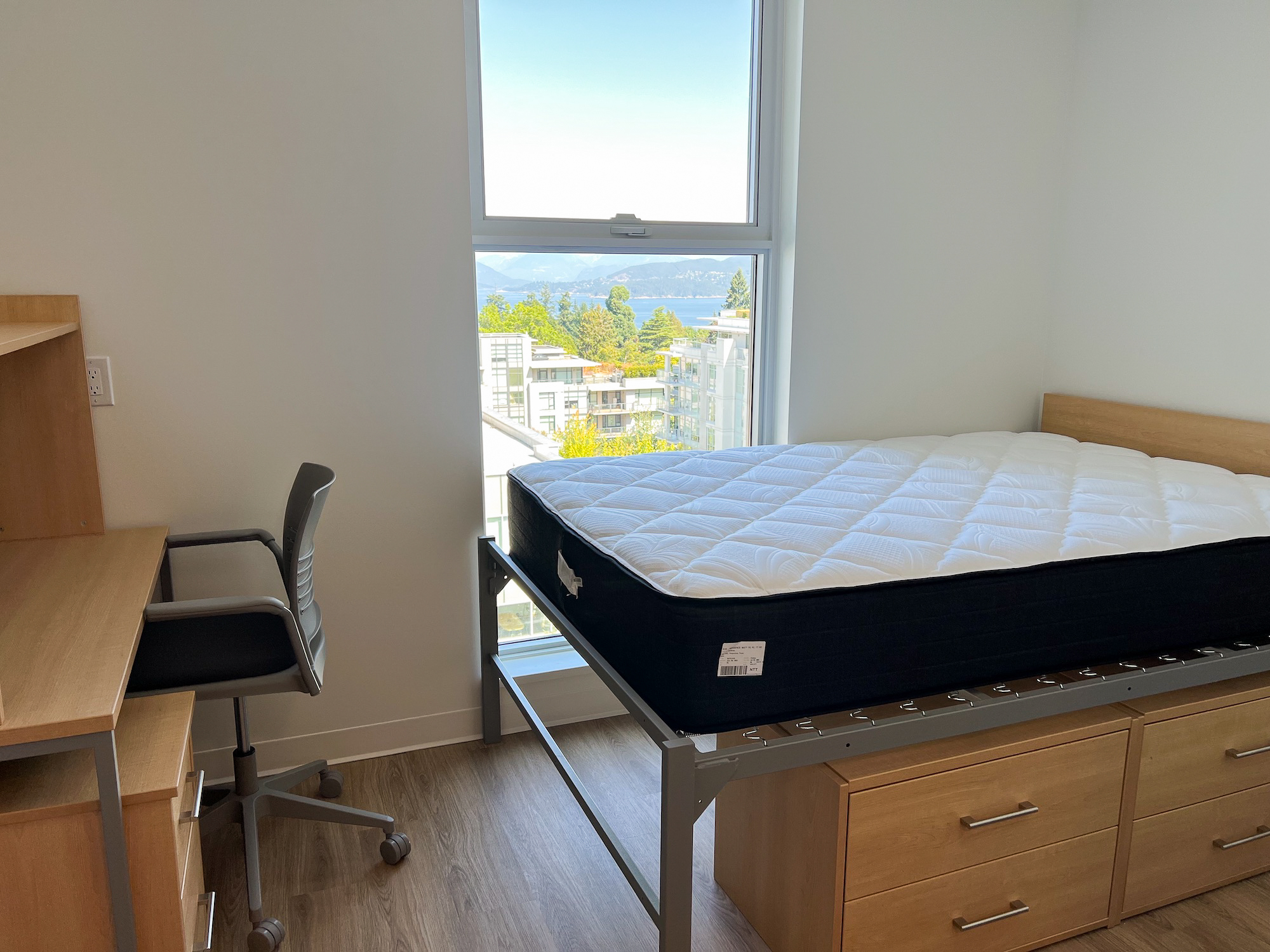 Interior of a room in the new Brock Commons North building. The room is on a higher floor that has an elevated bed with a row of drawers underneath on the right corner. On the left corner, there is a desk and a rolling hair. The window in the room overlooks a few other residential buildings and looks to the Burrard Inlet.