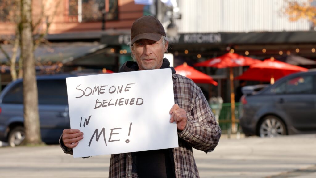 Homeless man named James holding up a handwritten sign that says Someone Believed In Me.