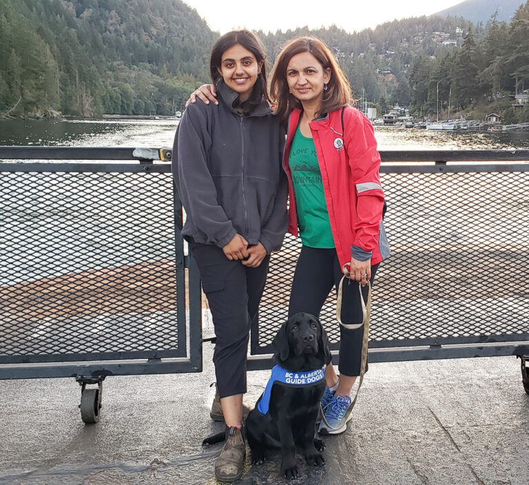 The mother-daughter duo, Jessie (on the right) and Sehaj (on the left) standing, while Daniel is sitting on the deck of a ferry, posing for the camera. Daniel is wearing a vest that says "BC & Alberta Guide Dogs"