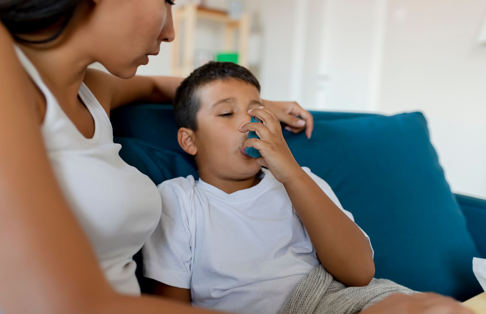 A child sits on a couch using an inhaler, accompanied by an adult who appears to be providing comfort and support.