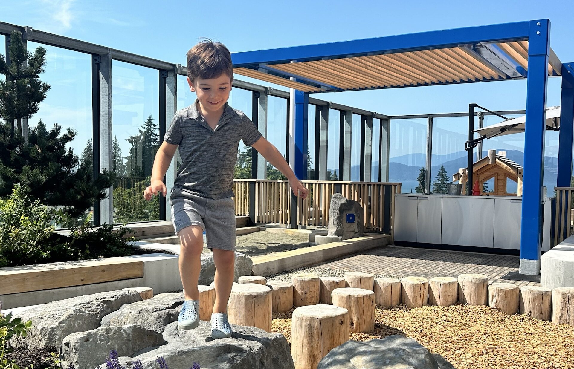 Child balancing on large wooden logs in an outdoor play area with a scenic mountainous backdrop.