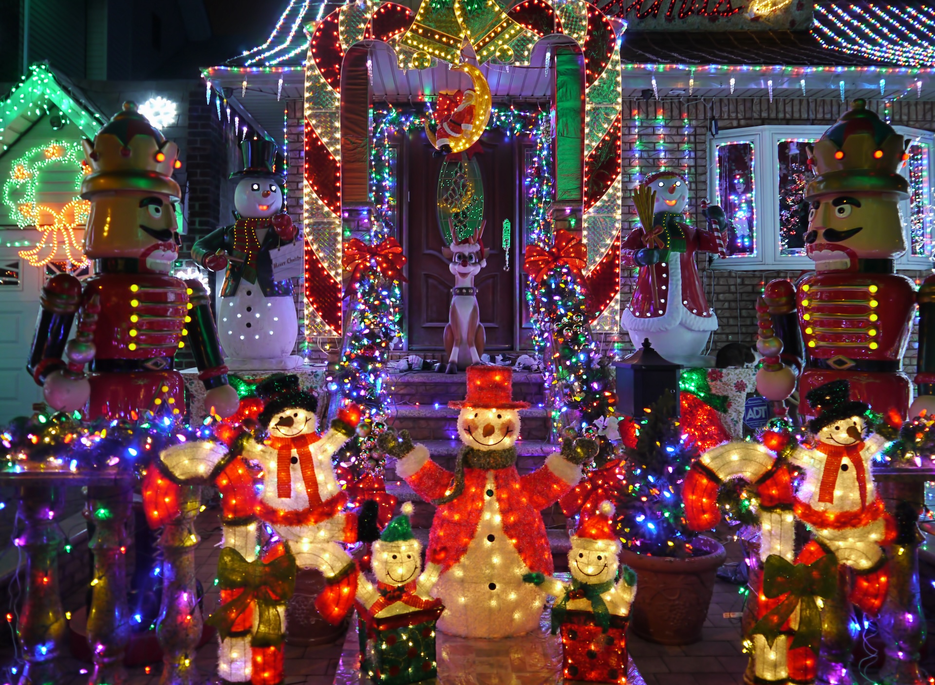 A brightly lit holiday scene featuring giant nutcrackers, snowmen, colorful lights, and festive decorations in front of a house. A reindeer stands on the porch surrounded by glowing ornaments.