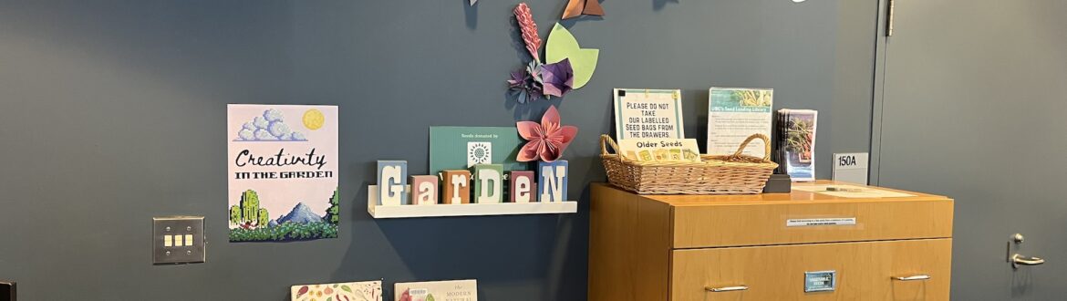 A Seed Lending Library display with a blue wall, origami decorations, a “Garden” sign, gardening books, and a wooden cabinet for seed storage.